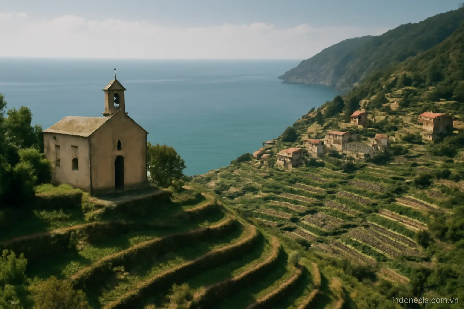 A hillside village with a small chapel and terraced fields descending to the sea, wide view, no text